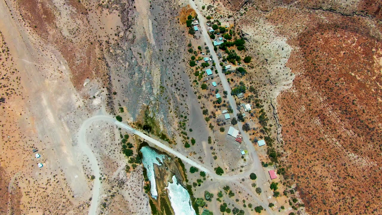 Top-down view over San Juanico, Baja California Sur with the bright water channel lower in frame, the dirt road crossing nearby, scattered buildings, trees, and no visible people.