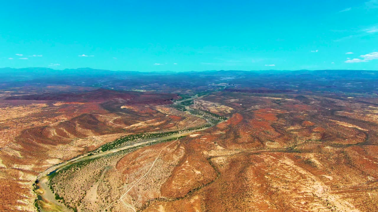 High aerial view of a river bend, dry hills, road traces, and distant mountains.