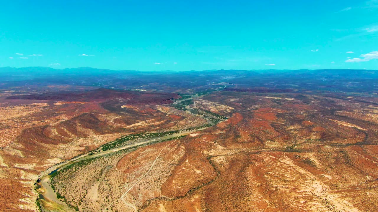 Mid-clip aerial frame over San Juanico, Baja California Sur, showing the river channel and road cutting through reddish arid hills toward a blue mountain horizon, with no visible people.