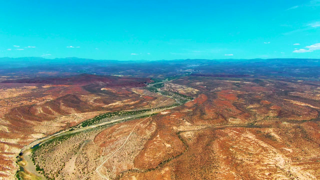 Wide aerial view near San Juanico, Baja California Sur showing a winding desert road beside a narrow river, red-brown hills, distant mountains, and no visible people.