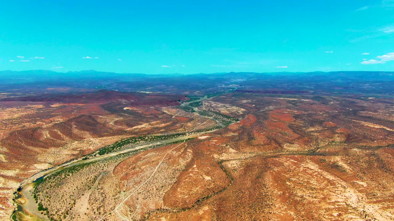 Wide aerial frame over San Juanico, Baja California Sur, showing sparse green vegetation along a curving river within red and tan arid hills, with no visible people.