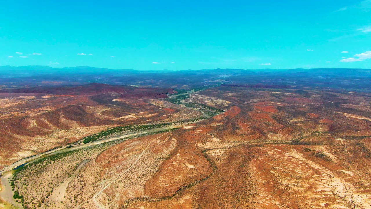 Aerial view near San Juanico, Baja California Sur, with red-brown desert hills, a winding pale road, distant mountains, and no visible people.