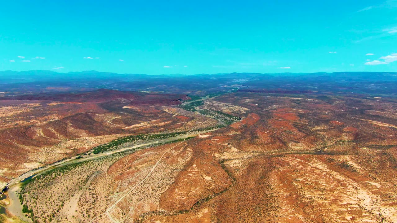 High aerial view near San Juanico with dry red hills in the foreground, a winding road through the midground, blue mountains in the distance, and no visible people.