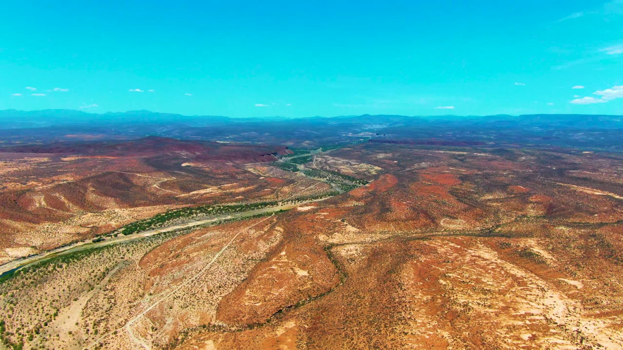 Opening aerial view near San Juanico, Baja California Sur, showing a road beside a green river corridor through red-brown desert hills with distant mountains and no visible people.