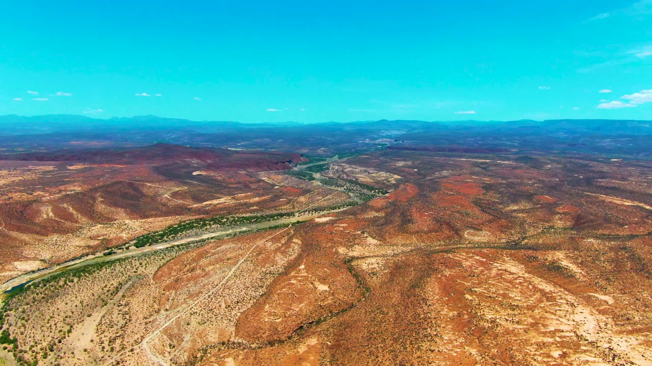 Third frame holds a high aerial composition of a winding road beside river greenery, pale desert flats, red hills, and distant mountains in San Juanico, with no visible people.