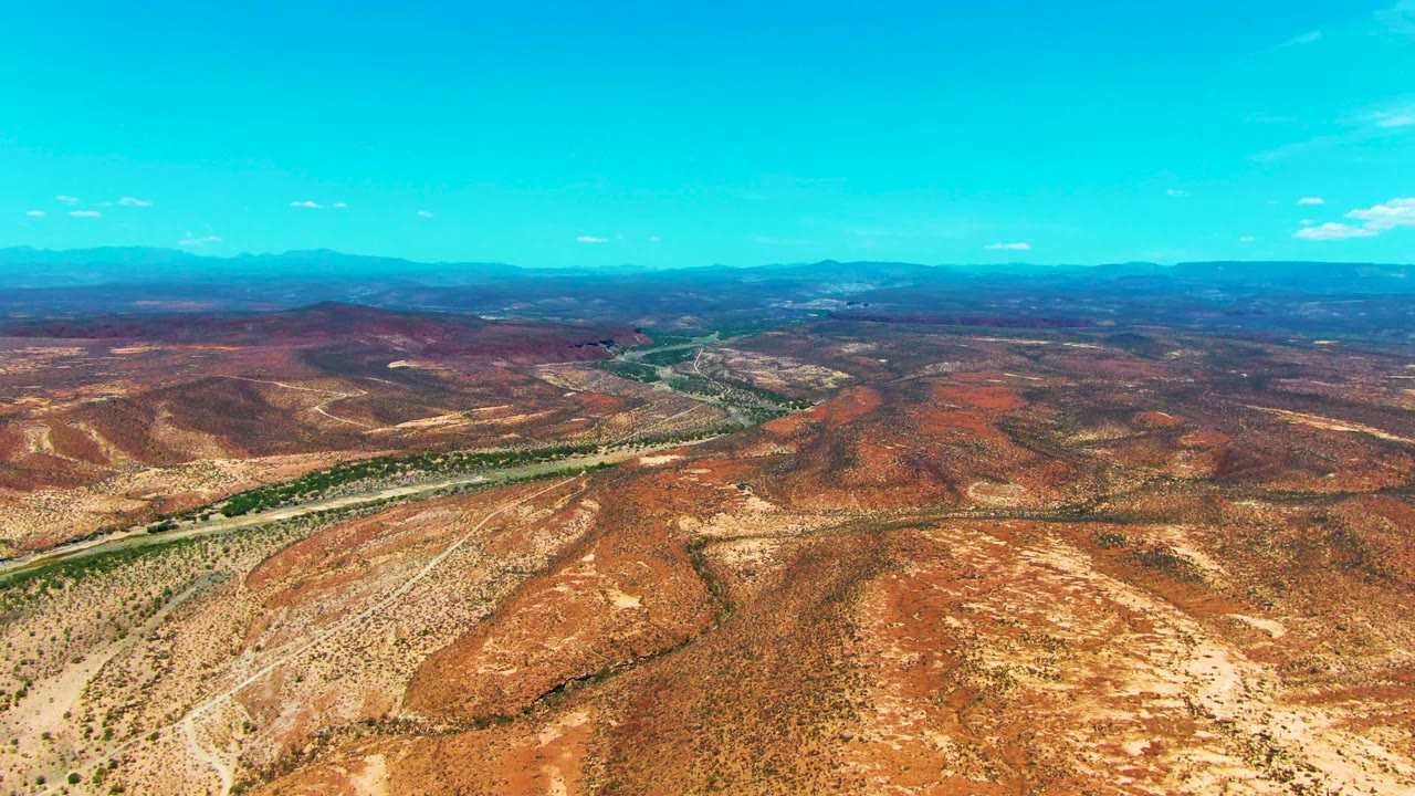 Aerial view over San Juanico, Baja California Sur, with a narrow river corridor cutting through red and tan desert hills, distant mountains, clear blue sky, and no visible people.