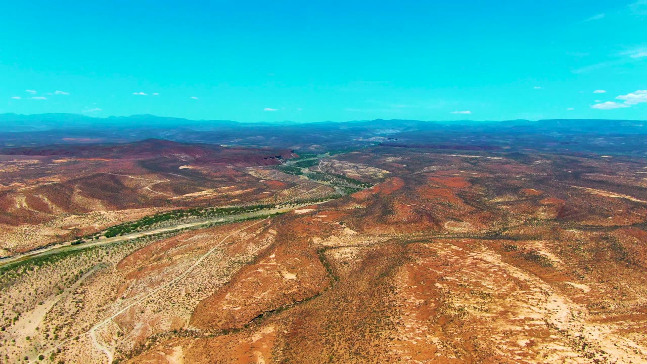 Wide aerial frame from San Juanico, Baja California Sur, showing sparse vegetation on dry red-brown terrain, a green river channel through the midground, distant mountains, and no visible people.