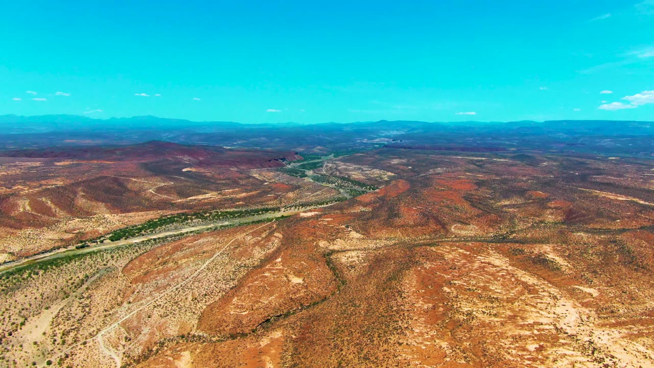 High aerial view in San Juanico, Baja California Sur, with arid red hills, pale exposed ground, a narrow green river band, hazy blue mountains, and no visible people.
