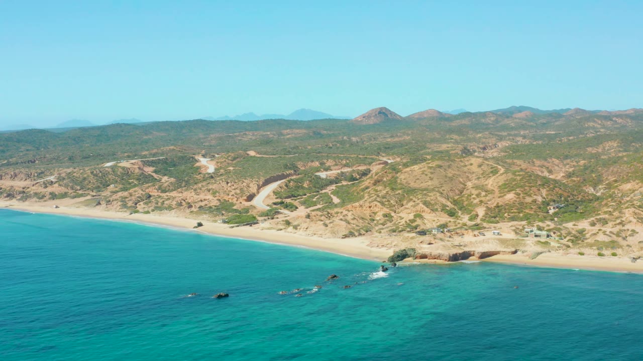 Aerial view at El Tule Los Cabos with turquoise ocean in the foreground, a pale sand beach, a small rocky point, dry green-brown hills, and no visible people.