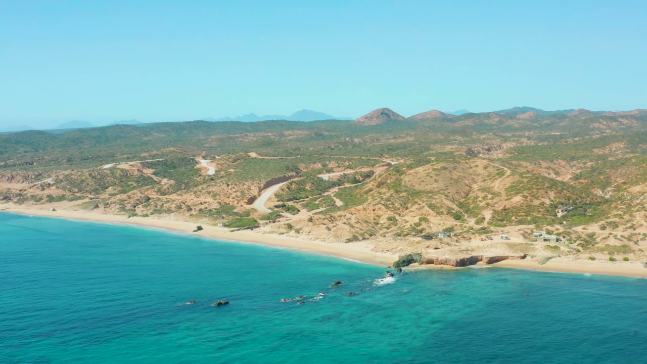 Wide aerial frame of El Tule Los Cabos showing blue-green water, a long sandy beach, scattered shoreline rocks, hillside tracks, distant hills, and no visible people.