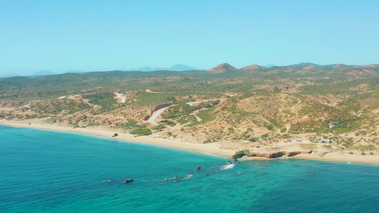 El Tule Los Cabos coastline viewed from above with turquoise sea, pale beach, a curving dirt road through dry hills, rocky shoreline details, and no visible people.