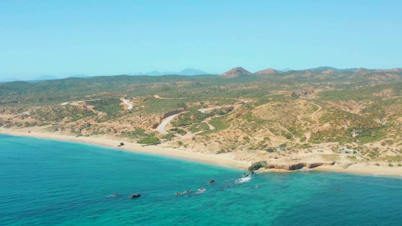 Aerial beach scene with clear water, small rocks offshore, and brown-green hills.
