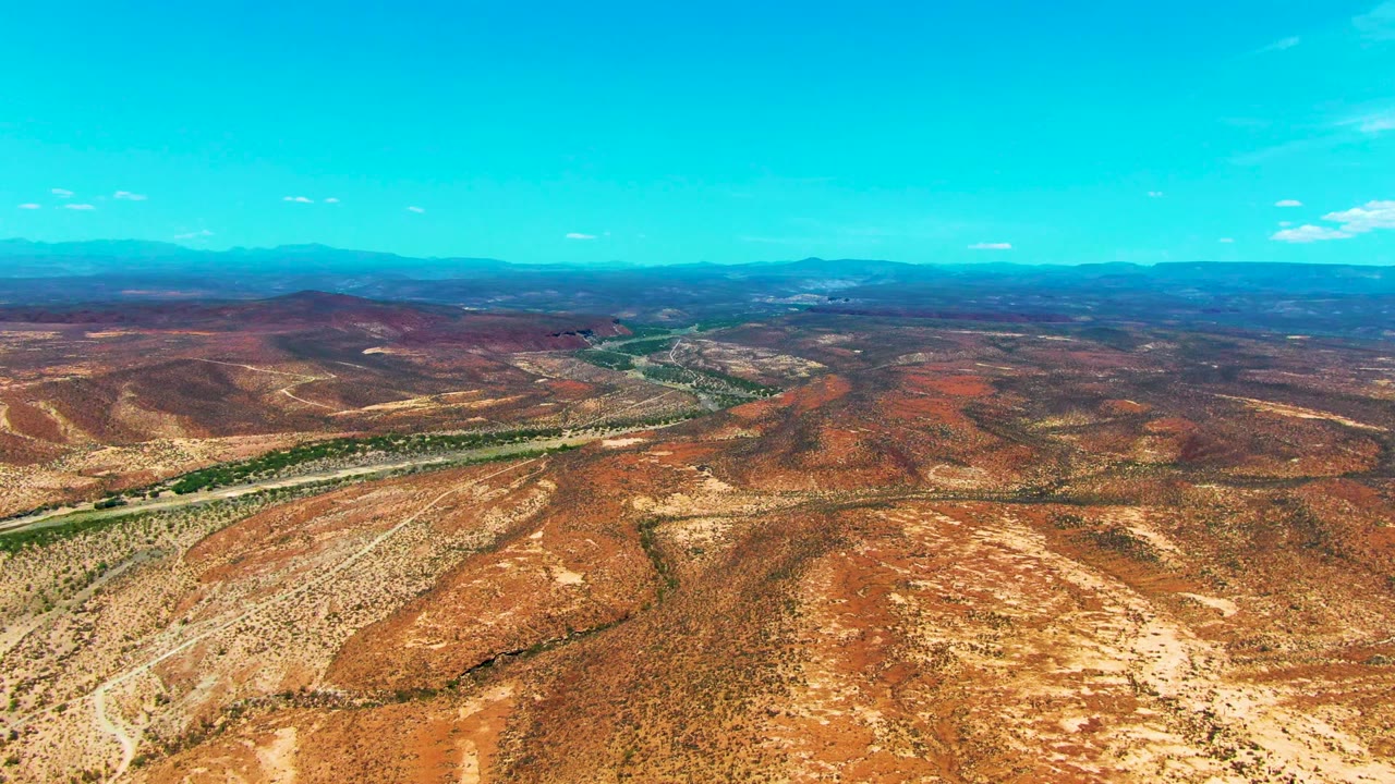 Opening aerial frame over San Juanico, Baja California Sur, showing red-brown desert terrain, a winding green river corridor, distant mountains, blue sky, and no visible people.