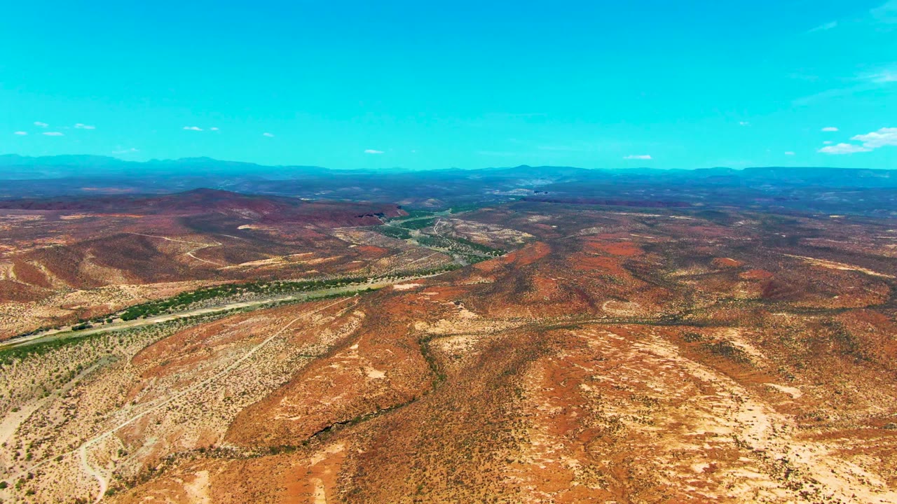 Mid-clip aerial view in San Juanico, Baja California Sur, looking over scrubby red-brown desert toward a winding green river corridor, a road line, distant mountains, and no visible people.
