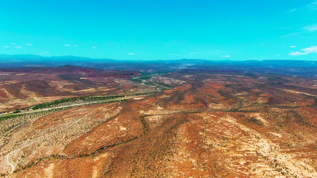 Wide aerial frame over San Juanico, Baja California Sur showing the road threading through arid red terrain toward blue distant ridges, with no visible people.