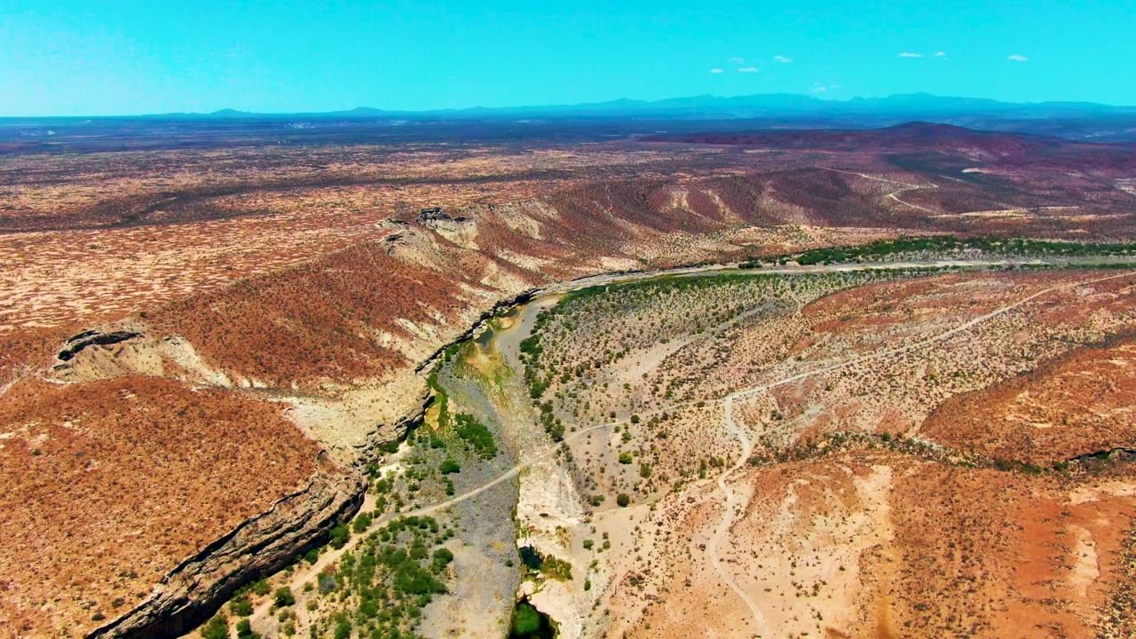 Aerial frame over San Juanico, Baja California Sur showing a winding river at lower left, red-brown desert slopes, sparse green vegetation, distant ridges, and no visible people.