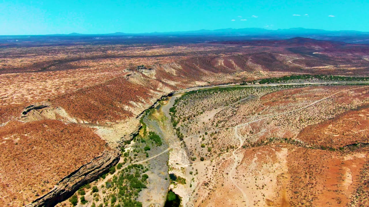 Drone aerial over San Juanico, Baja California Sur with the river bend more centered, scrub vegetation tracing the water, pale dry ground, layered hills, and no visible people.
