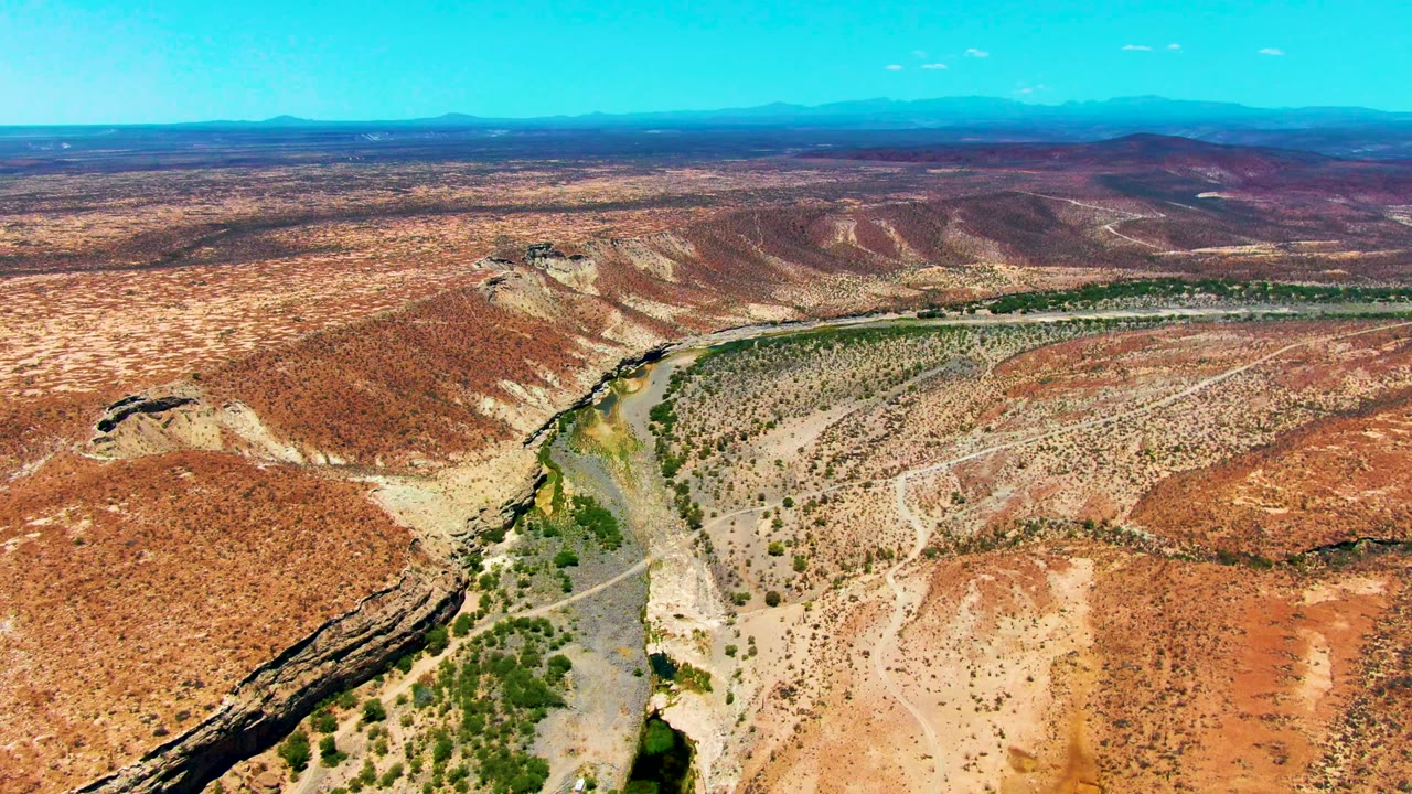 Wide aerial frame in San Juanico, Baja California Sur showing the river running diagonally through the lower frame, open red terrain, green channel vegetation, distant ridgelines, and no visible people.