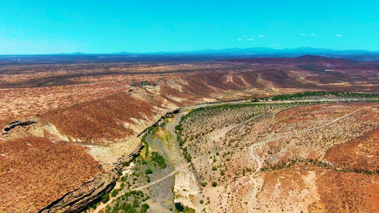 Opening aerial frame over San Juanico, Baja California Sur, showing a narrow river bordered by green vegetation, red-tan hills, dirt tracks, distant mountains, and no visible people.