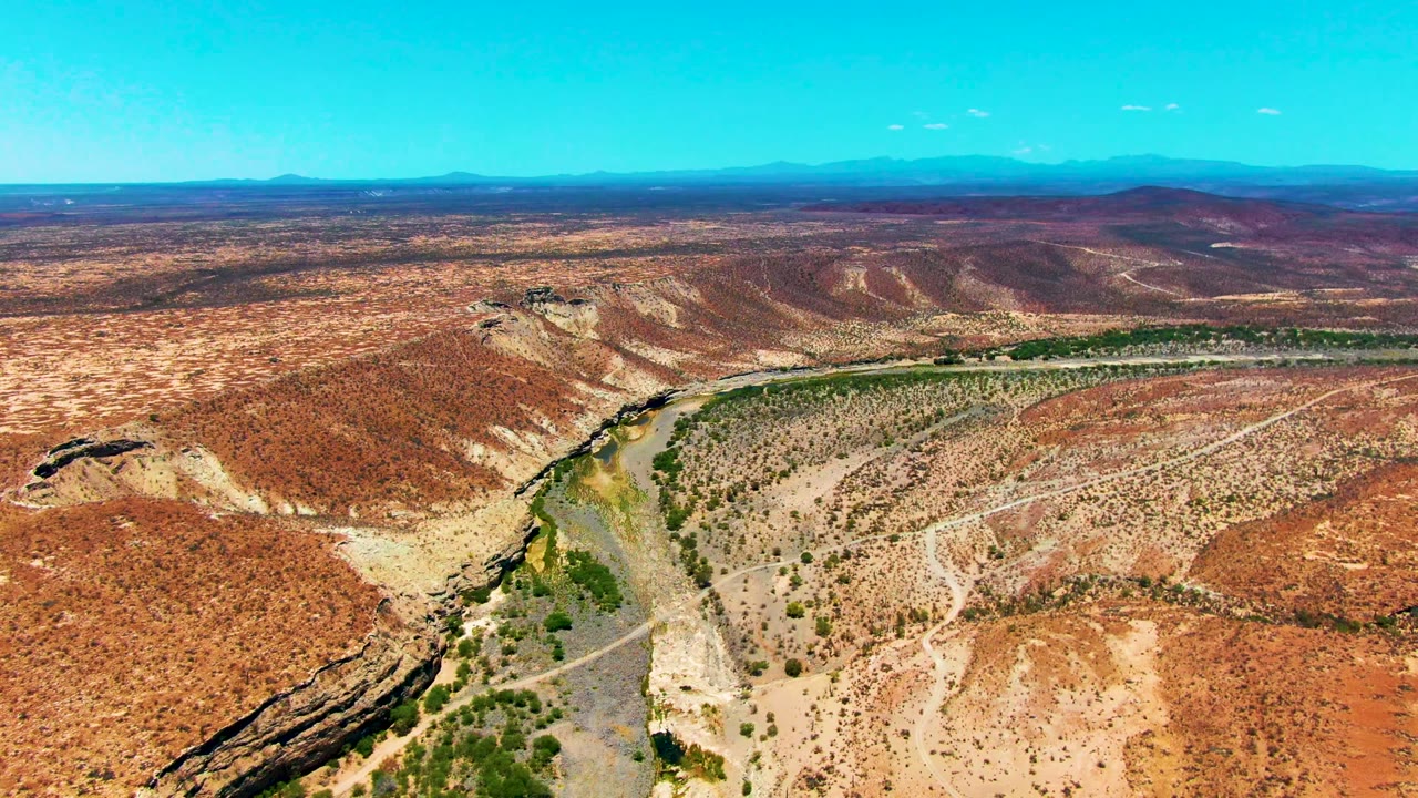 Third frame over San Juanico, Baja California Sur, showing the green river channel running across red and brown desert, with low ridges, faint roads, distant mountains, and no visible people.
