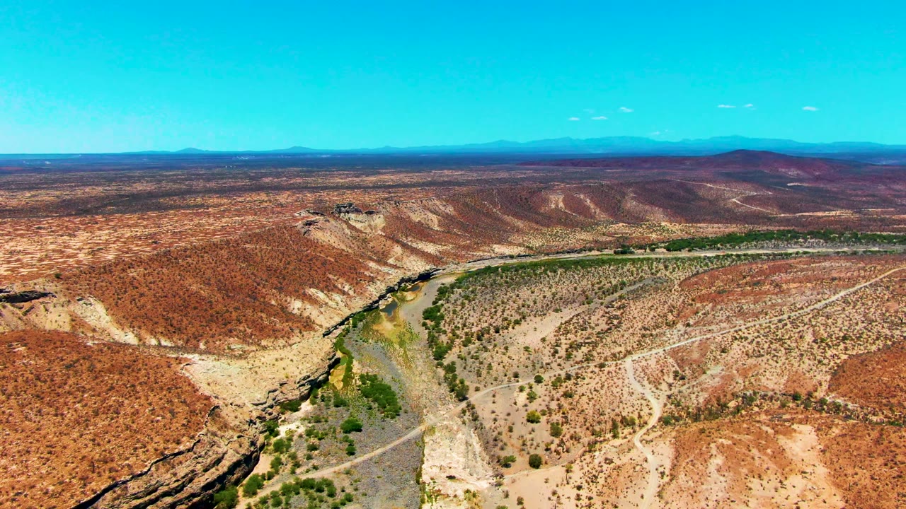 Aerial view in San Juanico, Baja California Sur, showing a narrow river, pale dirt road, red-brown desert hills, distant mountains, and no visible people at the start of the clip.
