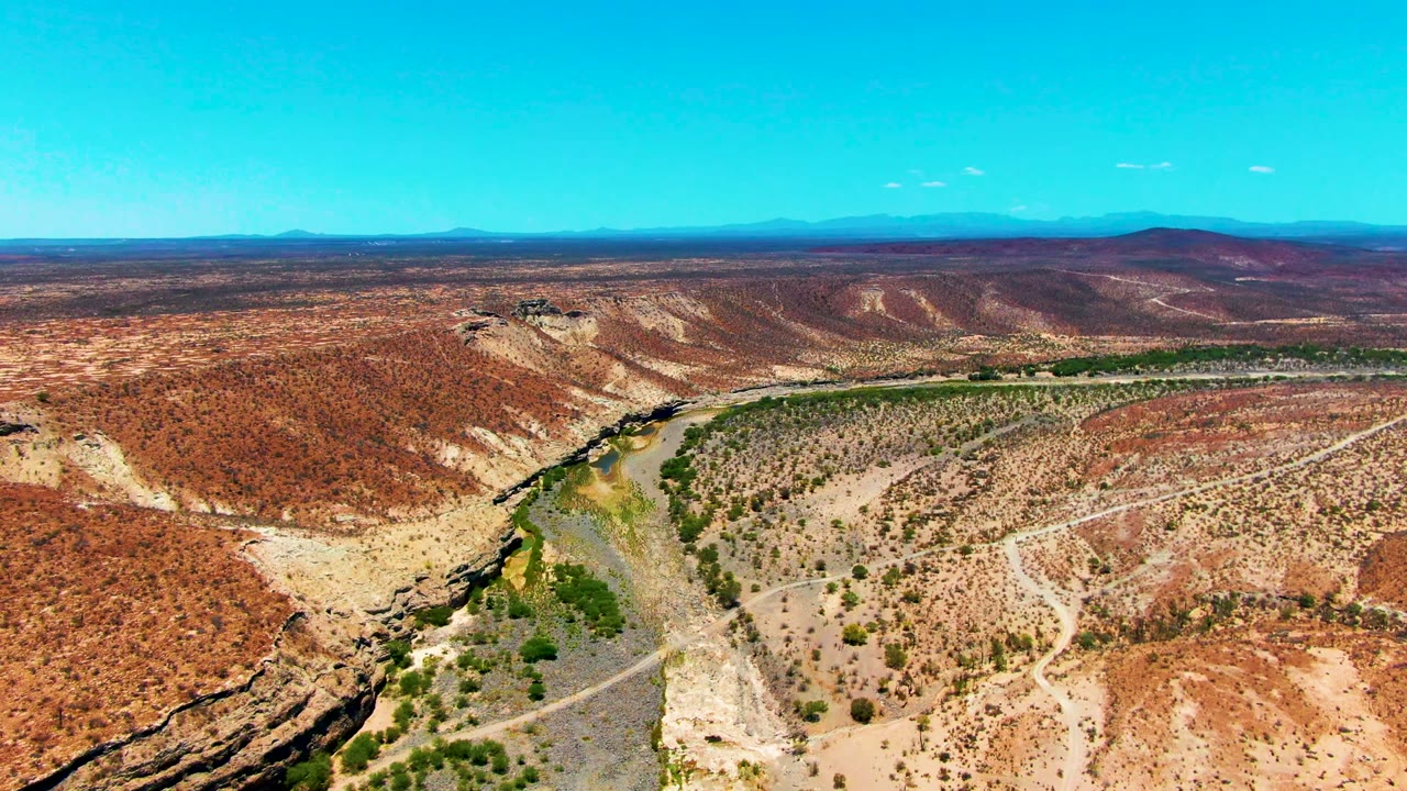 High aerial view over San Juanico, Baja California Sur, with a narrow river corridor, green banks, red-brown desert ridges, distant hills, clear blue sky, and no visible people.