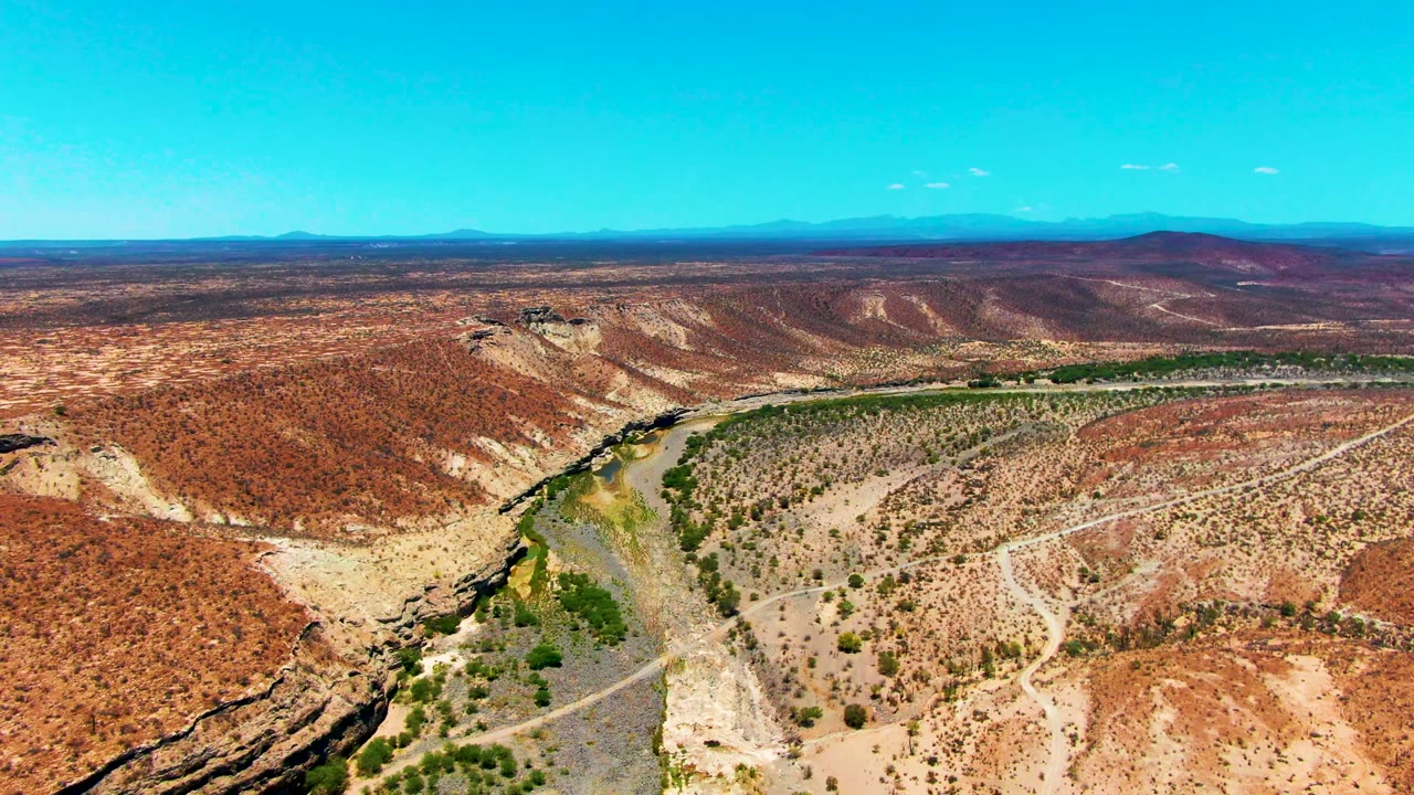 Wide overhead view of the San Juanico desert landscape with the river running through the left and center of frame, scattered scrub, pale dirt tracks, red-brown hills, and no visible people.