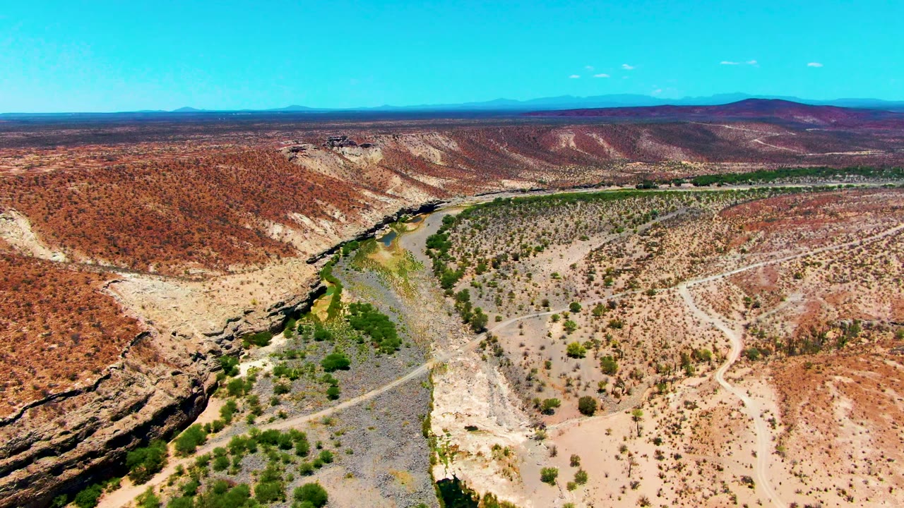 A wide aerial view over San Juanico, Baja California Sur shows a dry riverbed winding between red canyon slopes, green vegetation along the channel, distant mountains, and no visible people.