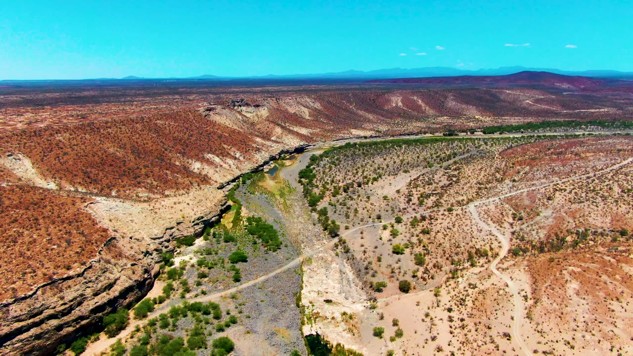 A broad aerial frame in San Juanico shows green vegetation tracing the dry wash below red and tan canyon walls, with a flat desert horizon and no visible people.