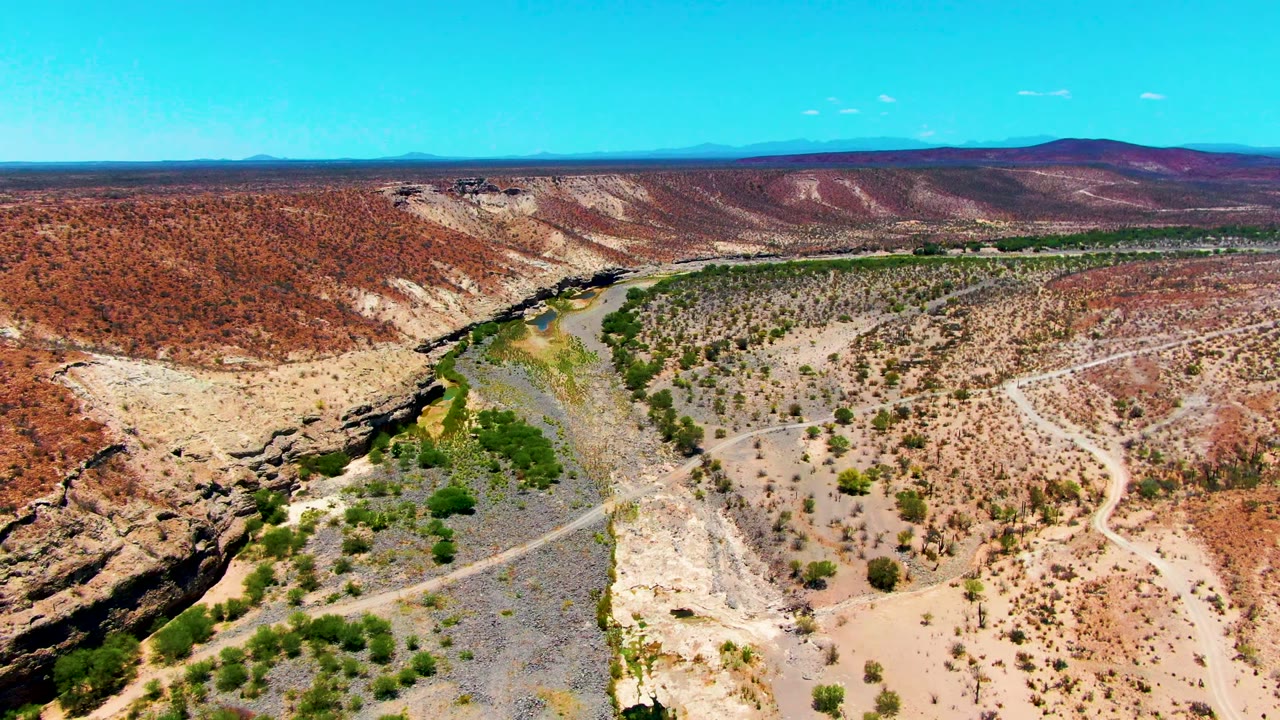 Aerial view near San Juanico, Baja California Sur, showing a winding river, pale canyon edge, red-brown hills, sparse green vegetation, and no visible people.