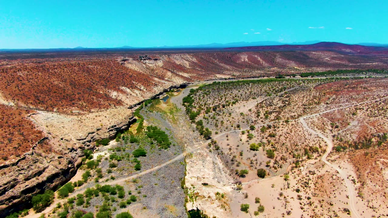 Drone view near San Juanico with rocky desert hills framing a green river corridor, pale eroded banks, a winding dirt track, blue horizon, and no visible people.
