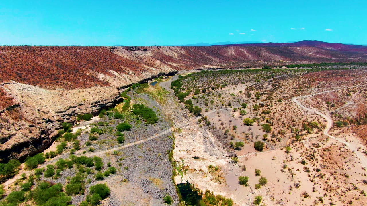 Aerial frame over San Juanico, Baja California Sur showing a dry riverbed road, sparse shrubs, red hills, distant mountains, blue sky, and no visible people.