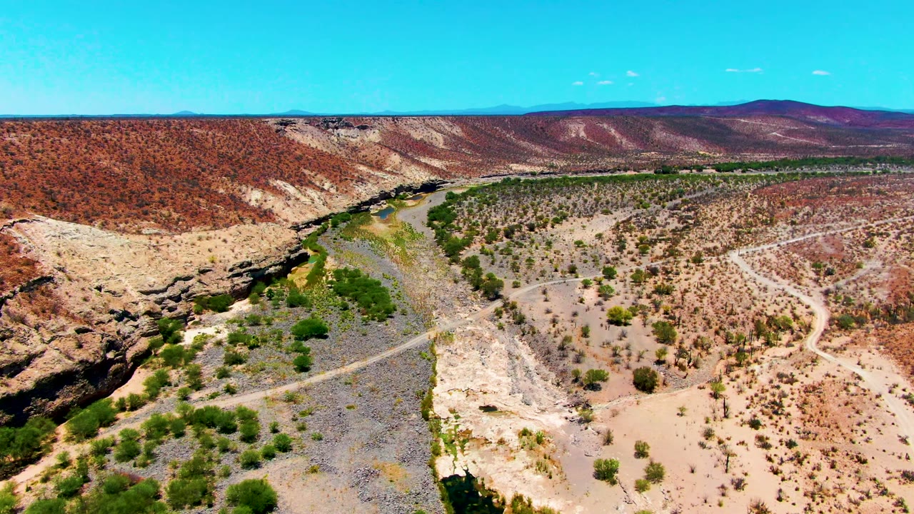 Aerial frame over the San Juanico desert terrain with a pale riverbed, scattered shrubs, reddish hills across the horizon, and no visible people.