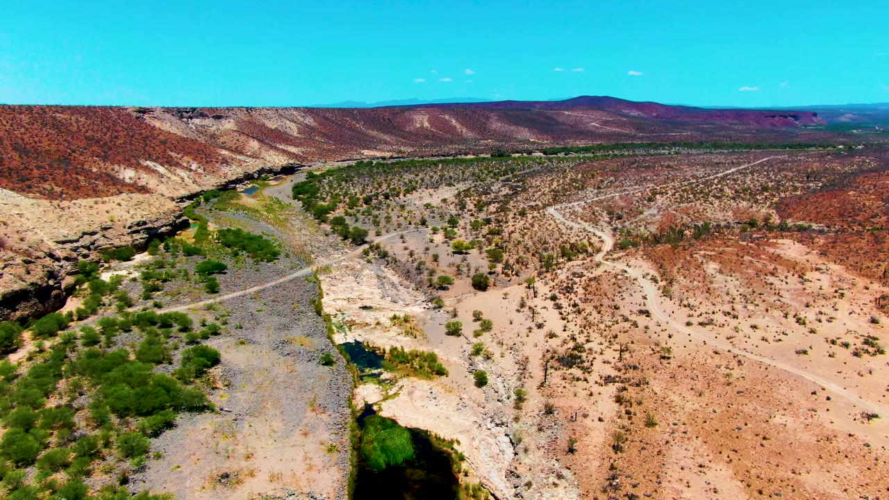 Opening aerial frame over a dry riverbed near San Juanico, Baja California Sur, with green shrubs beside shallow water, red hills beyond, and no visible people.