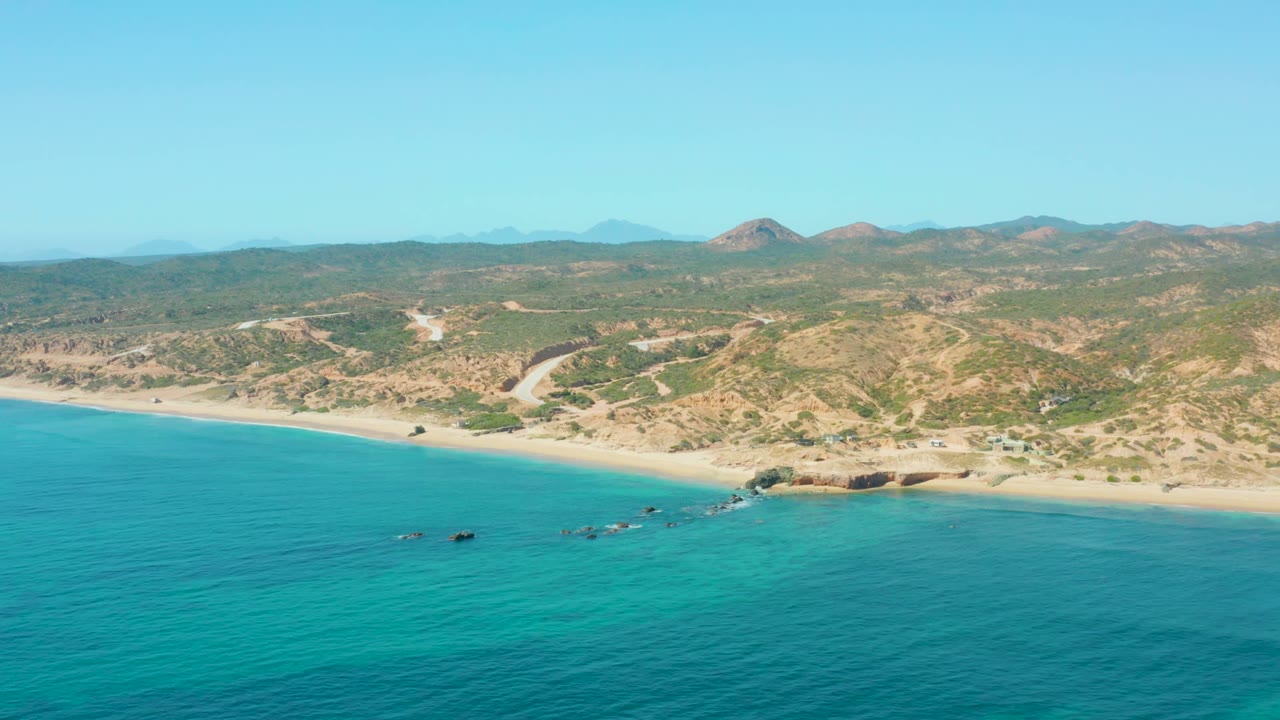 Aerial view of El Tule Los Cabos with turquoise ocean in the foreground, a pale sandy beach and rocky point along the shore, dry hills beyond, and no visible people.