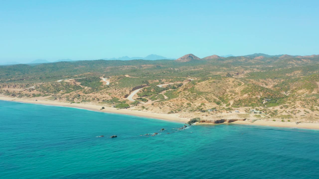 Aerial view of El Tule Los Cabos with turquoise water filling the lower frame, a sandy coastline and rocky shore detail at center, desert hills behind, and no visible people.