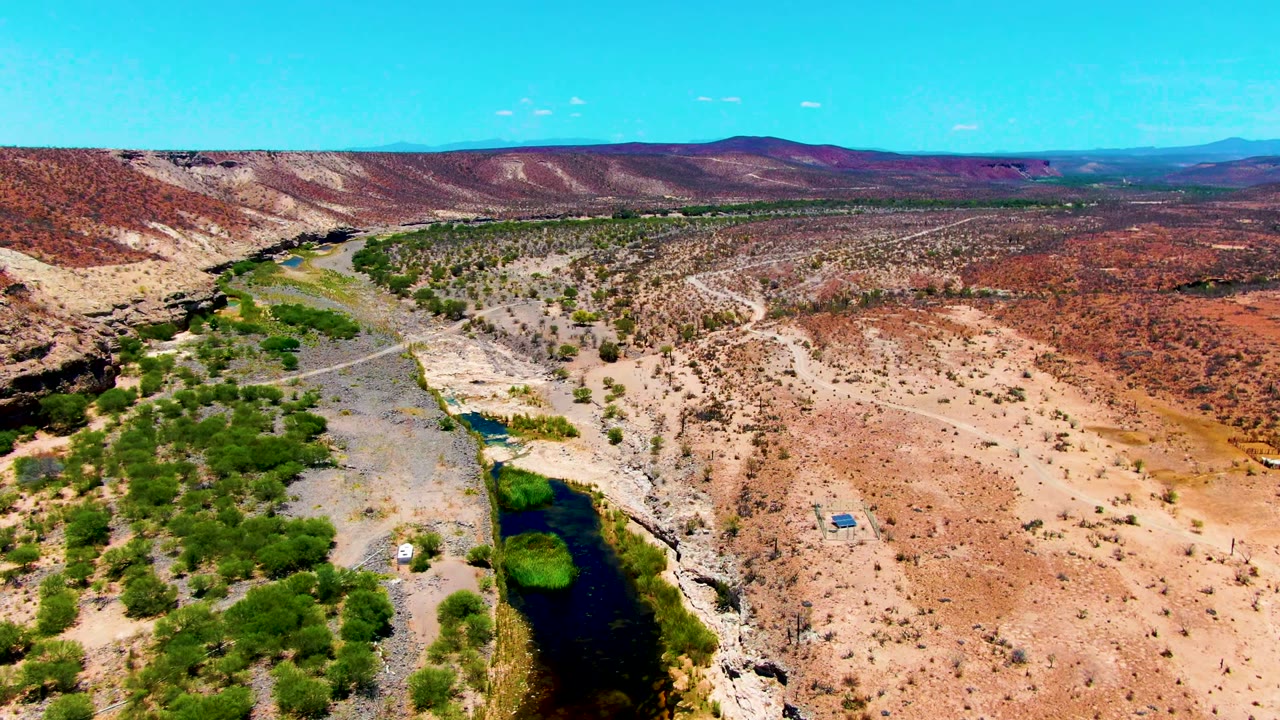 Aerial frame over San Juanico, Baja California Sur showing a dark river channel at lower left, pale gravel banks, red-brown desert slopes, dirt roads, distant hills, and no visible people.