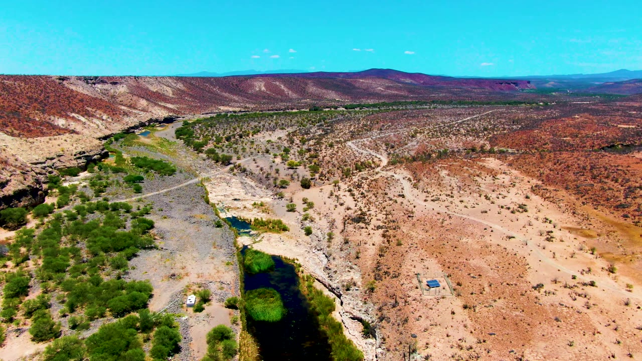 Wide drone-like frame over San Juanico, Baja California Sur showing the river tucked along the left side, scattered green vegetation, pale dirt roads, red desert terrain, a small vehicle near the lower area, and no visible people.