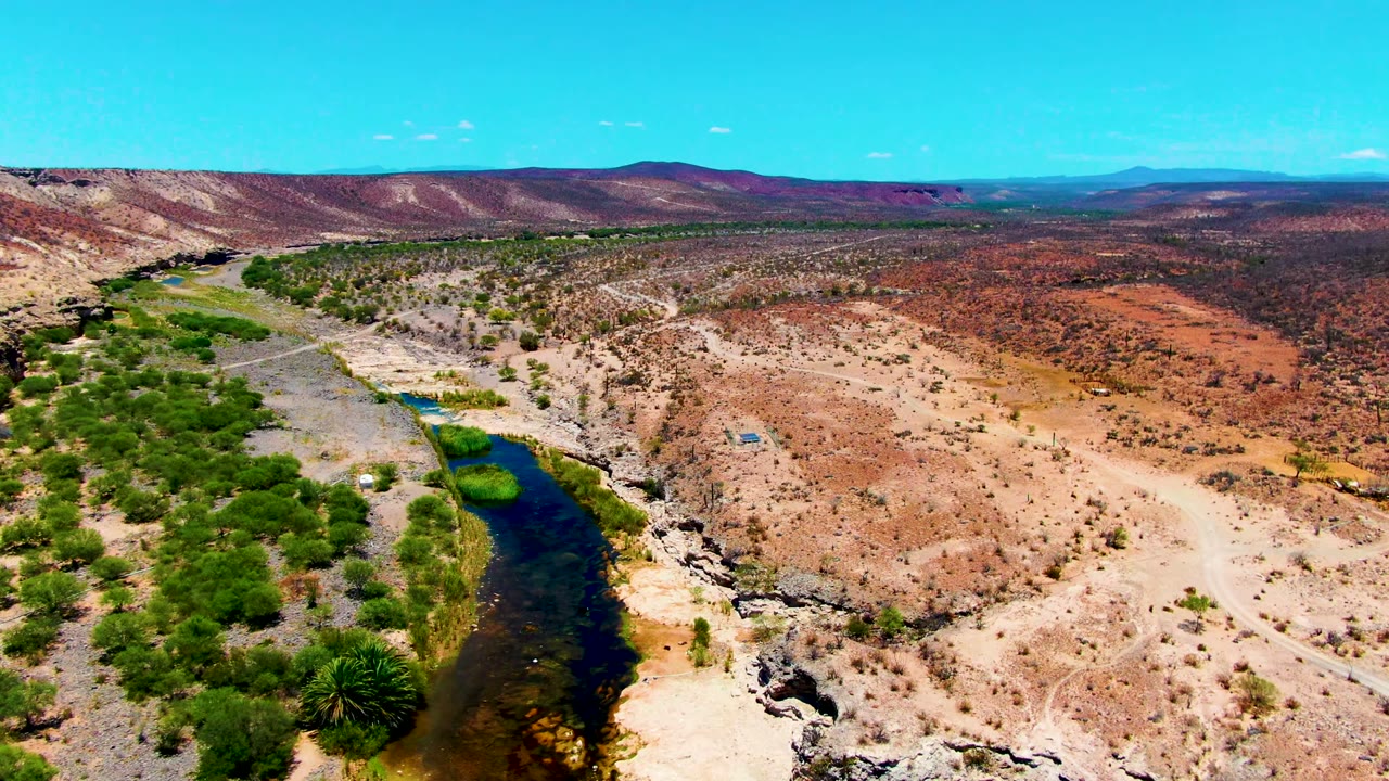 Aerial frame in San Juanico, Baja California Sur showing a dark river curving through rocky brown terrain with green trees along one bank and no visible people.