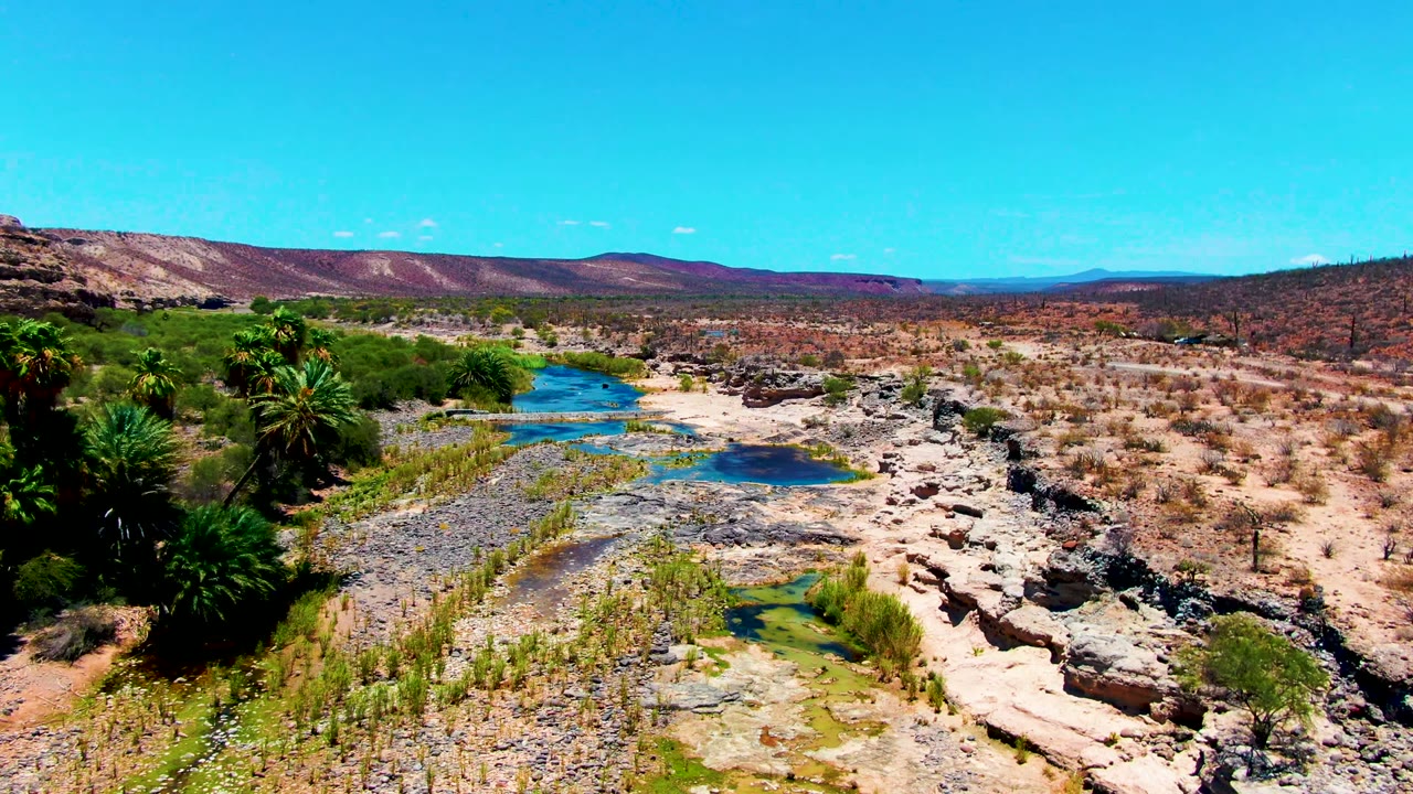 Aerial view in San Juanico, Baja California Sur showing a rocky dry riverbed with blue pools, palms at left, tan desert scrub, distant low hills, and no visible people.