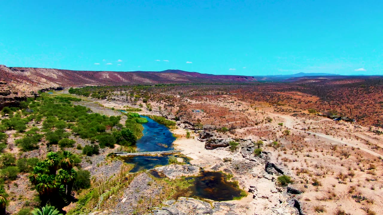 Wide aerial view in San Juanico, Baja California Sur, with a blue river bordered by green vegetation, rocky tan desert ground, low hills, and no visible people.