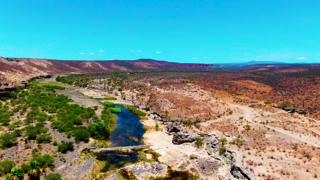 Aerial view over San Juanico with the river bending through green vegetation, a pale dirt road crossing dry ground, rocky outcrops, low hills, and no visible people.