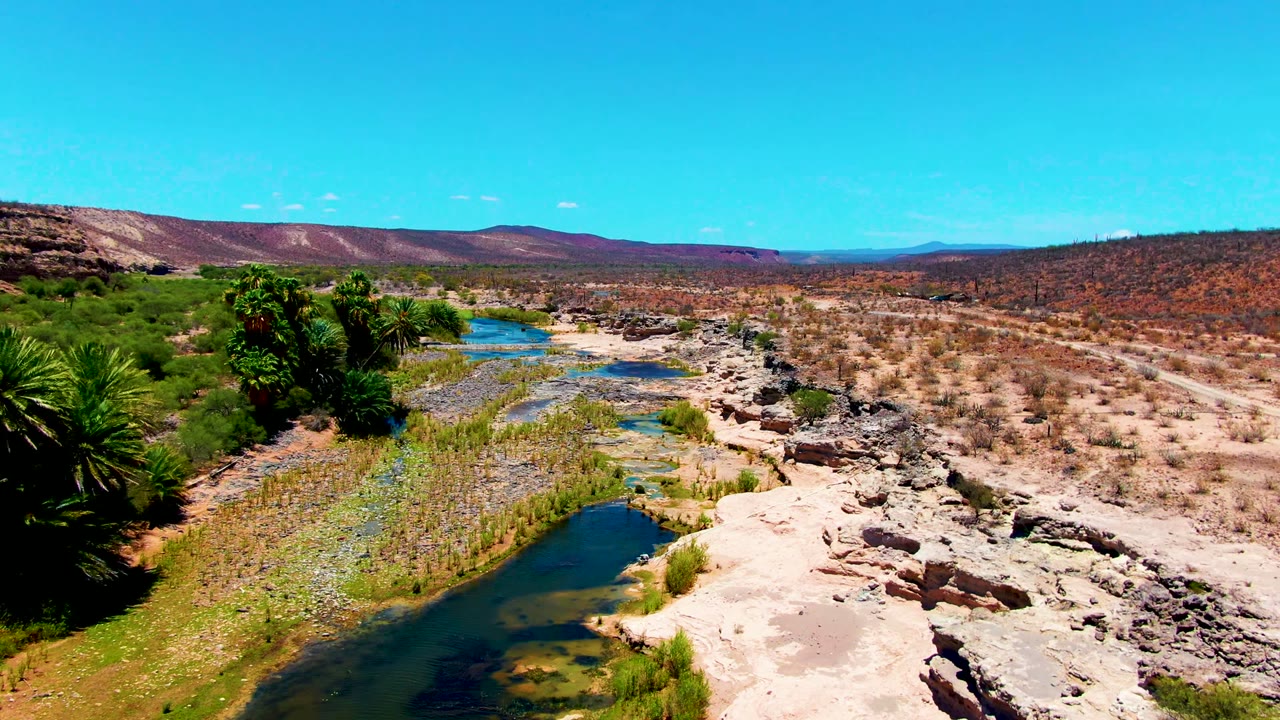 Wide aerial view over a shallow river channel, exposed rock, palms, scrub, and low hills in San Juanico, Baja California Sur, with no visible people.