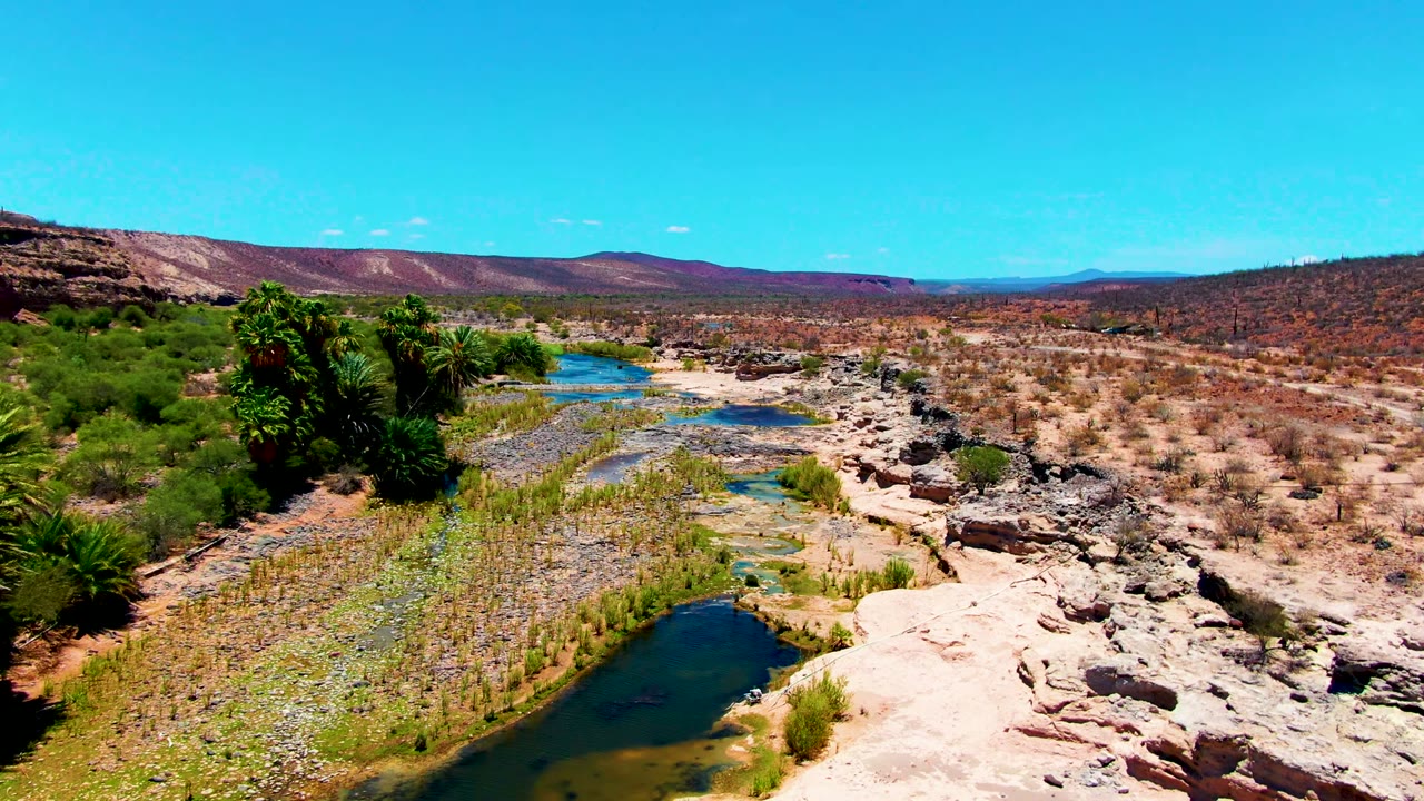 High wide view of a dry riverbed with narrow water patches, palms on the bank, scrubland, and low hills in San Juanico, Baja California Sur, with no visible people.