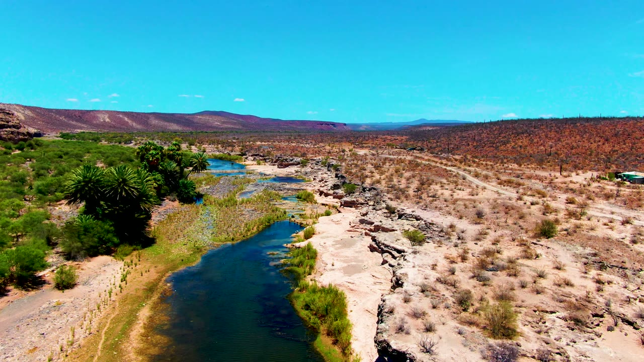 At 0:00, an aerial view in San Juanico, Baja California Sur shows blue-green river water, green bank vegetation, pale rock, dry scrub, distant hills, and no visible people.