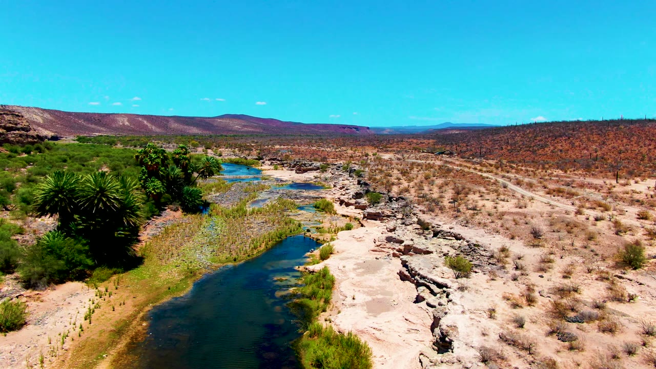 At 0:02, the frame shows the river running from the foreground into the desert, with rocky banks, scattered shrubs, a dirt road on the right side, distant hills, and no visible people.