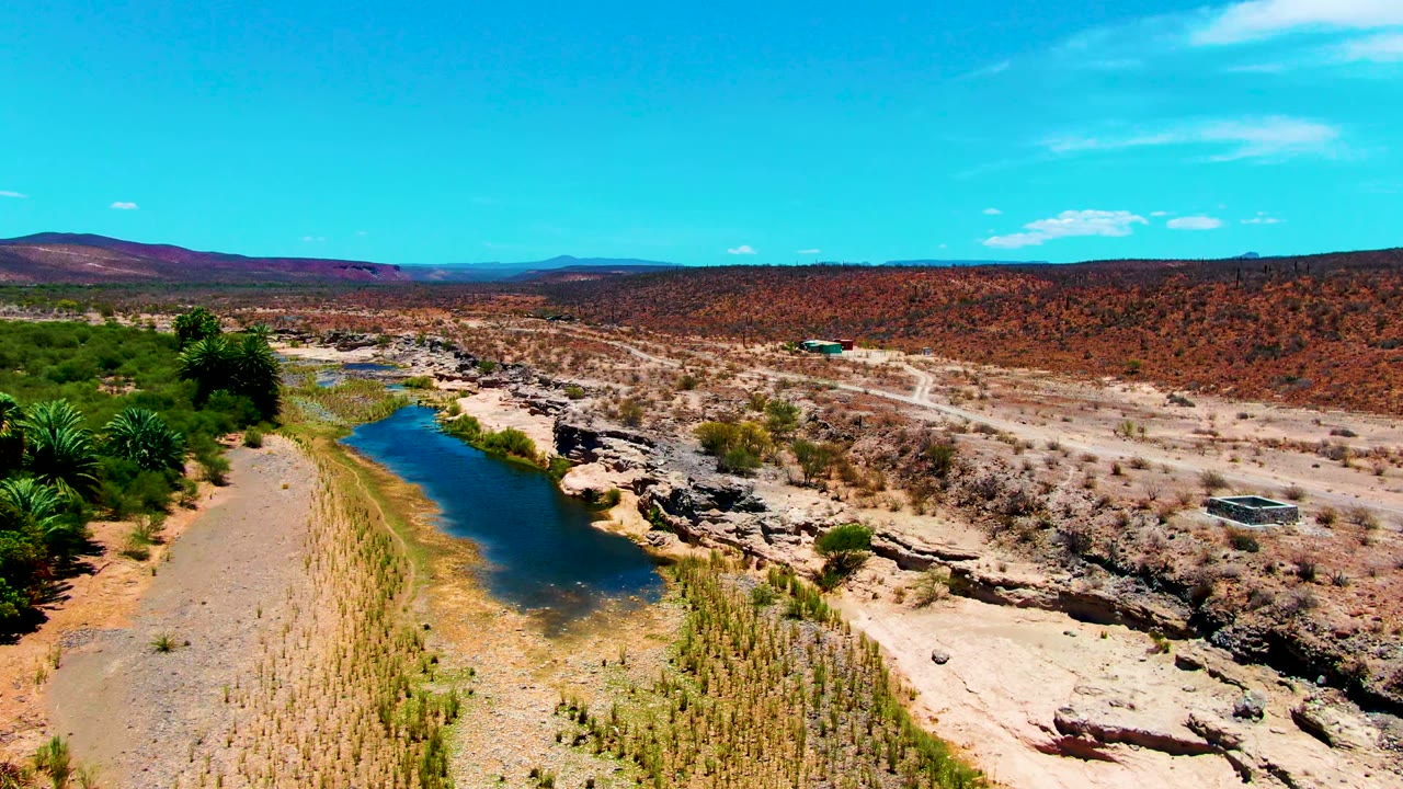 Aerial frame over San Juanico, Baja California Sur showing a blue-green river along pale sand, rocky banks, desert scrub, distant hills, and no visible people.