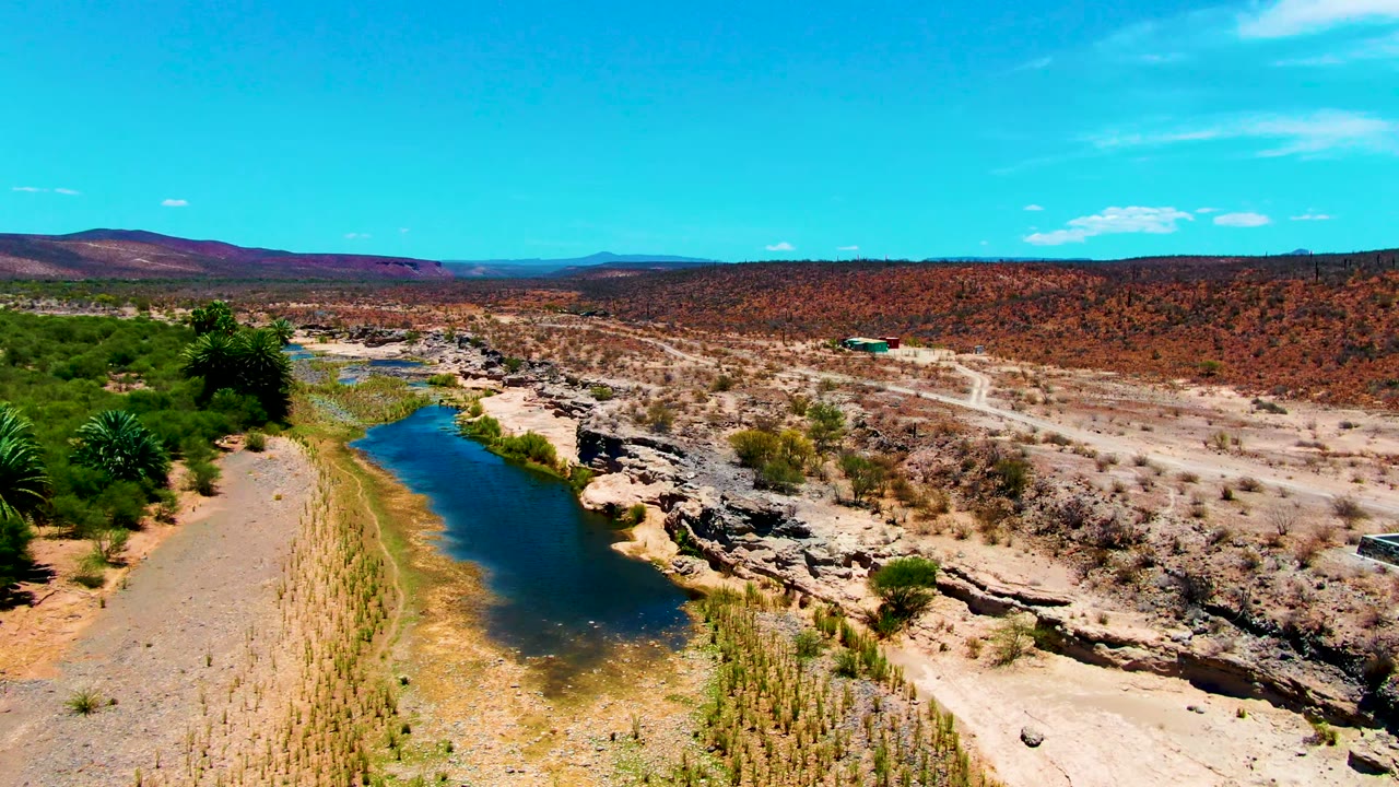 San Juanico aerial frame showing the river on the left, pale rock shelves and scrub in the center, a dirt track with a small vehicle to the right, and no visible people.