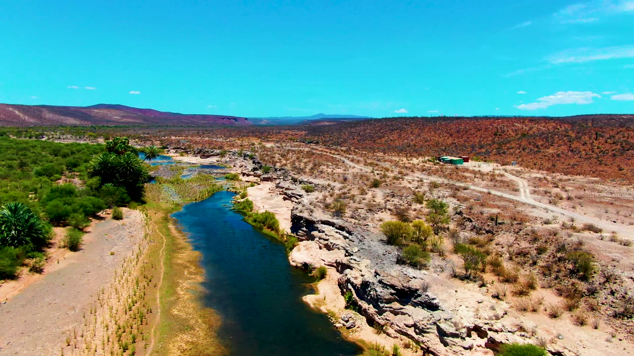 Opening aerial view in San Juanico, Baja California Sur, with a blue-green river at left, rocky tan banks, sparse desert vegetation, distant hills, and no visible people.