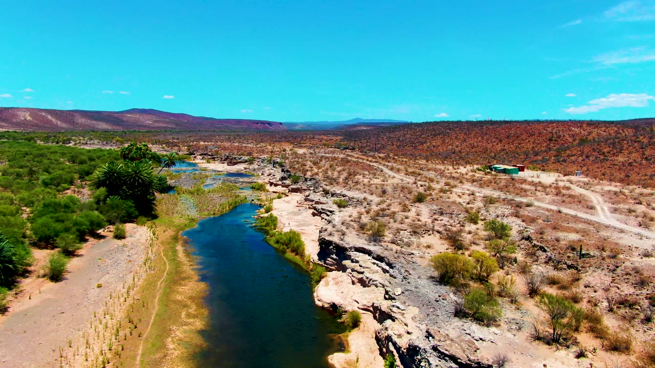 The San Juanico aerial view shows a river bend beside green vegetation on the left, exposed rocks on the right, open scrubland beyond, and no visible people.